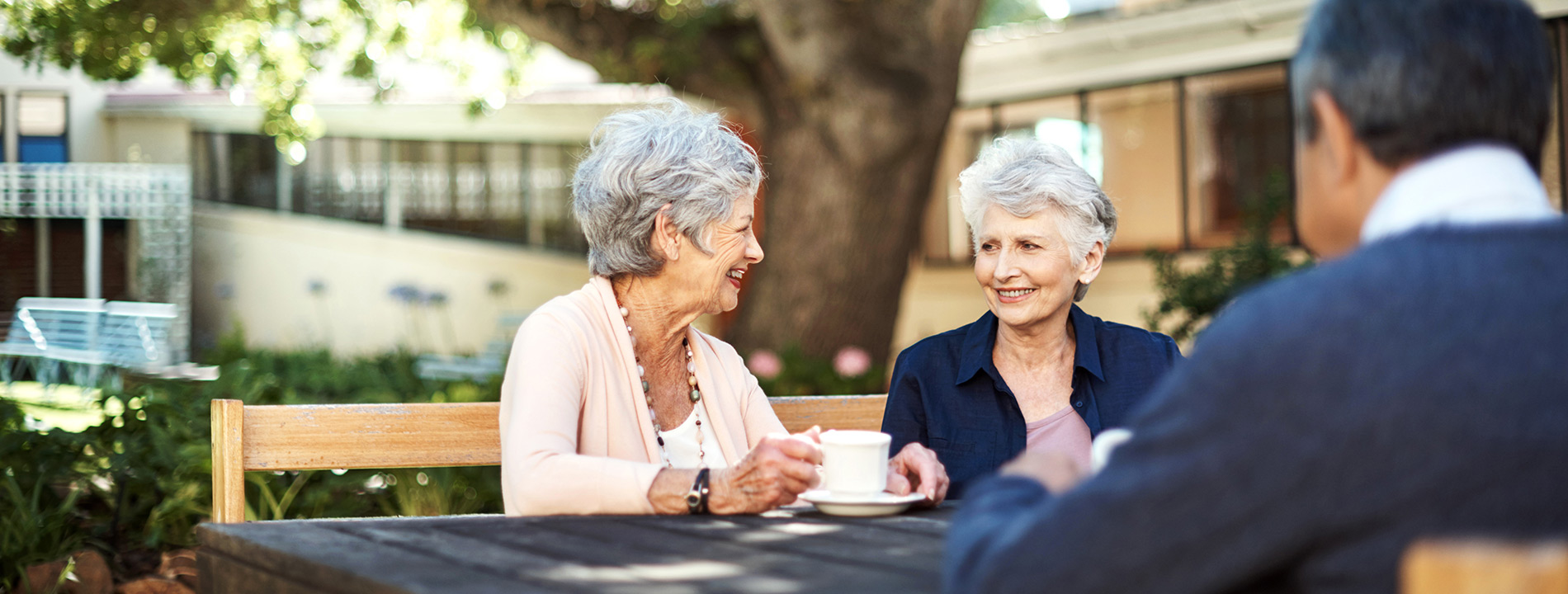 Two residents with coffee are sitting next to each other at a table smiling at each other. Another resident is sitting across from them. 